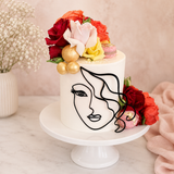 Cake with floral decorations and a face drawing on a white stand.